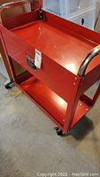 Red metal rolling shop cart with two shelves and chrome handles, shown from a side angle revealing wheels and overall shape.