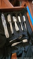 Photo showing a variety of flatware pieces including knives, forks, spoons, butter knife arranged on black cloth inside wooden drawer.