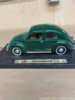 Green 1951 Volkswagen Beetle 1/18 scale model on black display stand with Maia branding plaque, left side view showing detailed body and part of beige interior.
