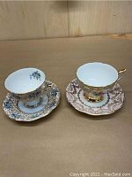Two Royal Standard bone china teacups with saucers, one set with blue floral pattern and gold accents, the other with gold floral pattern.