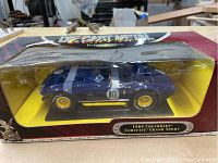 Front-side view of the diecast 1964 Chevrolet Corvette Grand Sport model inside the box showing dark blue car with yellow rims and white racing stripes in its original packaging.