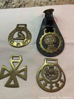 Four brass horse bridle medallions displayed on a white surface, showing a haystack/tree medallion on black leather, a cross-shaped medallion, a thistle flower horseshoe, and a sitting cat medallion.