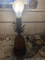 Full view of the lit lamp showing the pineapple brass base and lampshade on a countertop.
