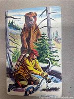 Front view of illustrated bear trap card depicting man in yellow and red outdoor gear setting a chain trap in a forested snowy landscape.