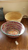 Two woven baskets placed on wooden floor: a natural straw basket with leather handles and a colorful round fabric basket.