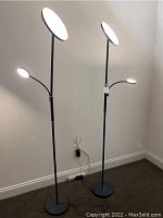 Two black metal floor lamps with tall poles, large round LED lights at the top, and smaller adjustable reading lights attached below. Both lamps are illuminated and plugged in.