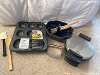 Wide shot of waffle maker, baking pans, brushes, wooden spoon, spatula, and pastry cutter arranged on floor in front of white fabric background.