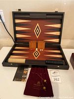 Backgammon board opened, showing inlaid wood design, wooden dice cups, checkers, dice, and branded velvet pouch on table.