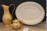 Photo showing the gold-toned metal pitcher, matching lidded teapot, and Lenox Halah cream china tray with gold-colored trim and embossed Hebrew text.