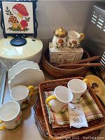 Wide view of lot including ceramic mushroom mugs, sugar and creamer set, wooden salad bowl, vintage mushroom trivet, napkin holder, wicker basket, plates, and spoon rest