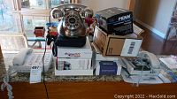 Lot of amplified phones and electronic boxes arranged on a counter, including a silver novelty rotary dial phone, multiple white amplified phones, and factory sealed electronic equipment boxes.