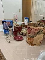 Wide view of all items including the waste basket, Conair hot lather machine box, ceramic soap and matching cup and dish, all placed on a bathroom counter.