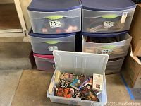 Seven stacked plastic storage bins with one open bin showing assorted books inside.