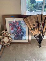 Room corner showing a framed floral artwork, large Asian folding fan, and decorative wall clock grouped together.