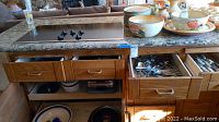 Photo shows wooden cabinet with five drawers and open lower shelf containing plates and cookware. Top granite countertop with black electric stovetop and stacked ceramic bowls and plates. Two drawers open displaying metal flatware.