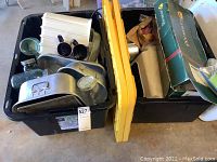 Two black plastic bins filled with various kitchenware including pans, jars and mugs, and packed with a yellow bin lid visible.