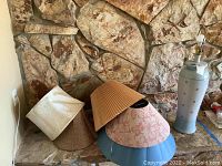Photo of ceramic lamp base and multiple cloth lampshades arranged on floor against stone wall.