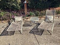 Two outdoor loungers with woven metal frames and patterned cushions seen from the front, with one small round table between them, placed on a stone patio near a garden.