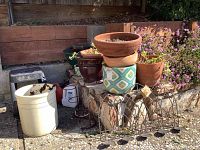 Outdoor scene showing multiple ceramic pots stacked and arranged on a stone ledge and ground. Featured are a large white ceramic crock, several terra cotta pots, a geometric glazed pot, and a metal candle votive with six candles slots in front.