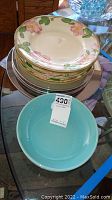 Stack of floral rimmed plates and turquoise bowl with seller price tag on glass table