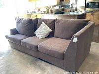 Brown microfiber three-seat sofa with loose back cushions placed in kitchen area. Shows fabric wear and slight discoloration.
