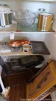 Photo of a shelf containing a large clear glass serving bowl, a gold metal tin, two large glass jars, and a Toastmaster griddle in its original box below the shelf along with various baking pans and utensils.