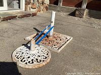 Three heavy metal umbrella bases arranged on concrete ground showing decorative scroll design, vertical poles, and attached blue straps.