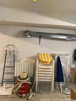 Shows stacked white plastic chairs, lounger with yellow striped cushion, foot stool, two blue umbrellas mounted on stands, vintage aluminum step ladder against wall, Milwaukee vacuum on floor.
