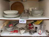 Shelf with various ceramic dishes, woven tray, glass salad bowls, metal graters, and kitchen utensils arranged.