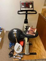 Full view of the exercise bike with accessories around it including dumbbells, exercise ball, hand weights, tennis racket, and operation manual on seat.