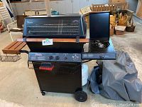 Front view of the Fiesta BBQ grill showing the main grilling compartment, wooden shelves on the left, side burner on the right with gray cover beside the grill, propane tank below, and wheels.