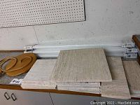 Stack of square beige textured floor tiles and white packaging tubes placed on a surface with a wooden tree swing nearby.