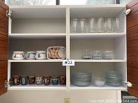 Glass cabinet showing clear glassware on the right side top and middle shelves; white ceramic bowls and small plates on the bottom right shelf; pottery cups on the bottom left and a few smaller cups with prints on the left middle shelf; and large conch shells in a plastic container on the middle left shelf.