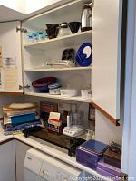 Photo of kitchen cabinet corner showing nesting mixing bowls in blue and red, clear glass casserole dishes, blue napkins or fabric, and electric hot plate sitting on countertop above white dishwasher.