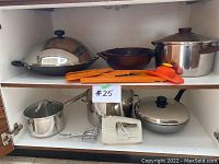 Wide view of kitchen cabinet showing assortment of kitchen pots, lids, a white Black & Decker electric hand mixer with metal beaters, and orange cooking utensils.