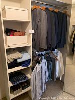 Wide view of closet shelving and hanging area showing folded sweaters, gloves, scarves, two white storage boxes, button-up shirts and assorted pants and shorts below.