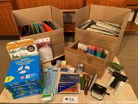 Wide shot of the stationery lot showing multiple cardboard boxes filled with binders, paper packs, markers, pencil crayons, various office supplies including scissors, staplers and hole punchers.