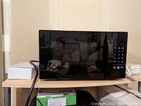 Front view of the black Amazon Alexa enabled microwave on a shelf with power cord and box beside it