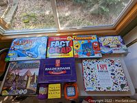 Photo of 9 boxed games lined up in sunlight on a window sill: Sorry Sliders, Fact or Crap, Connect 4, Fire of the Ice Lab, 7 Wonders, Balderdash, I Spy, The Tangrams Box puzzle, and Pank!