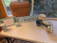 Photo showing full lot displayed on table: wicker basket, 'It's A Wonderful Life' sign, tin bucket, small green pot, photo frame, glass bowl with decorative eggs