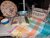 Overview of the lot showing the colorful checkered tablecloth, vintage-style Roman numeral clock, large metal garden container, two beige slipcovers, clear glass vase, small porcelain container, and decorative bowls.