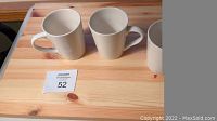 Three new white ceramic mugs with curved handles shown on wooden table surface.