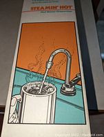 Close-up of box front showing Steamin' Hot hot water dispenser illustration with hot water flowing into cup.