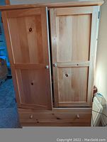 Front view of pine wood armoire showing two paneled doors and lower drawer with round wooden knobs. Left door slightly ajar due to incomplete closure.