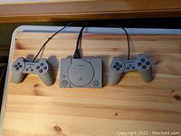 View of Sony PlayStation Classic console with two wired controllers on a wooden surface, showing full items included in the lot.
