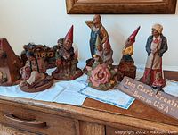 Multiple Tom Clark sculptures including gnomes, women, and men figurines displayed on table along with wooden signs including 'Born in USA' sign.
