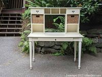 White wooden desk with a removable hutch placed outdoors against stone steps and greenery. The desk shows a keyhole on the large drawer below the desk surface. The hutch has two small drawers with metal label holders, two open cubby compartments, and two wicker baskets. The desk has four round turned legs.
