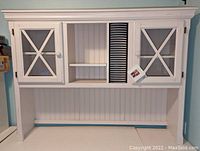 Front view of the white wooden hutch showing two glass paneled cabinet doors with cross decoration, open shelves, and built-in CD holder filled with CDs.