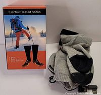 Electric heated socks grey color with black heel and toe areas, connected via wire, shown with packaging box illustrating winter hiking use.