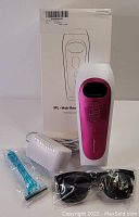Pink IPL hair removal device with white buttons, a separate white accessory head sealed in plastic, a blue razor in plastic wrapping, black protective glasses, and user guide booklet.